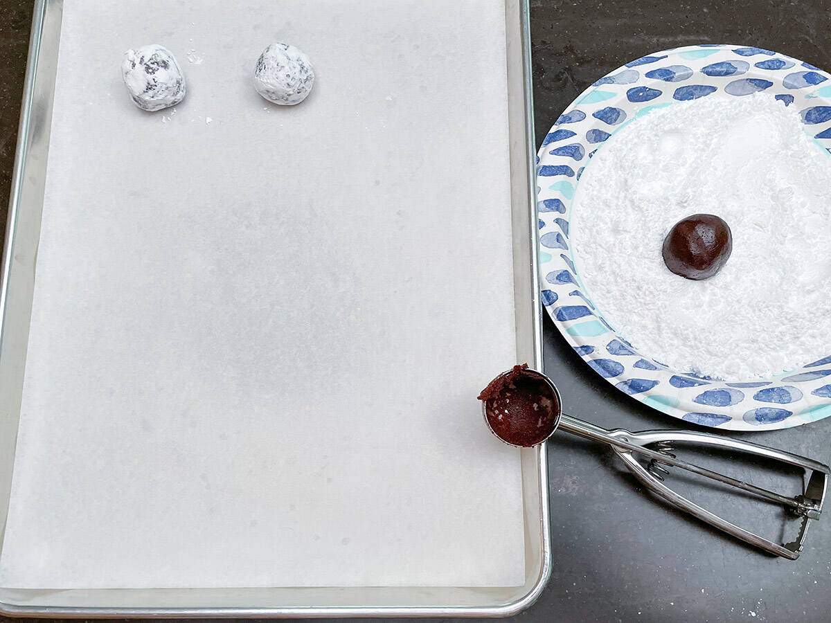 Scooping cookie dough and making into a ball then rolling in powdered sugar before placing on a sheet pan.