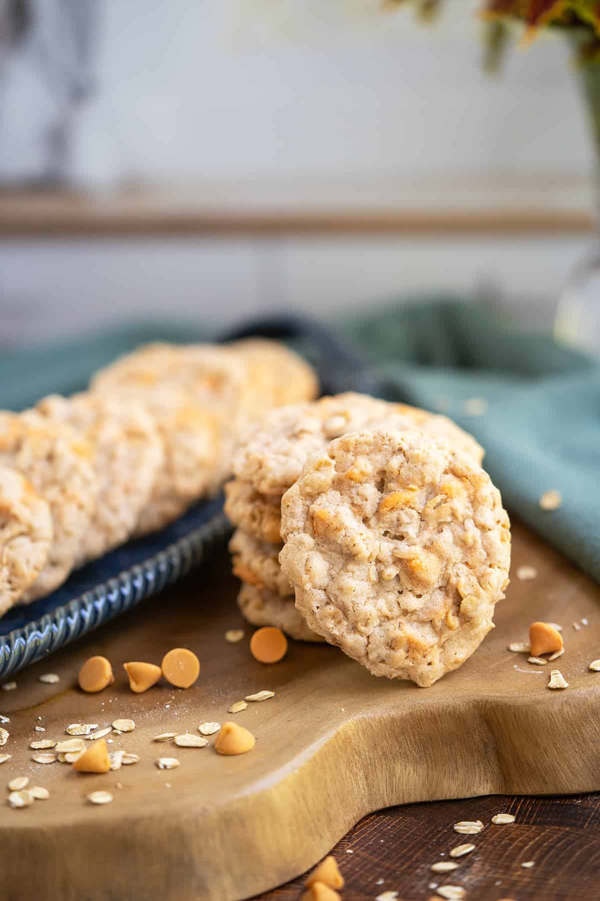 Stack of Old Fashion Oatmeal Scotchie Cookies on a wooden plank with butterscotch bits and oatmeal lying beside the cookies.