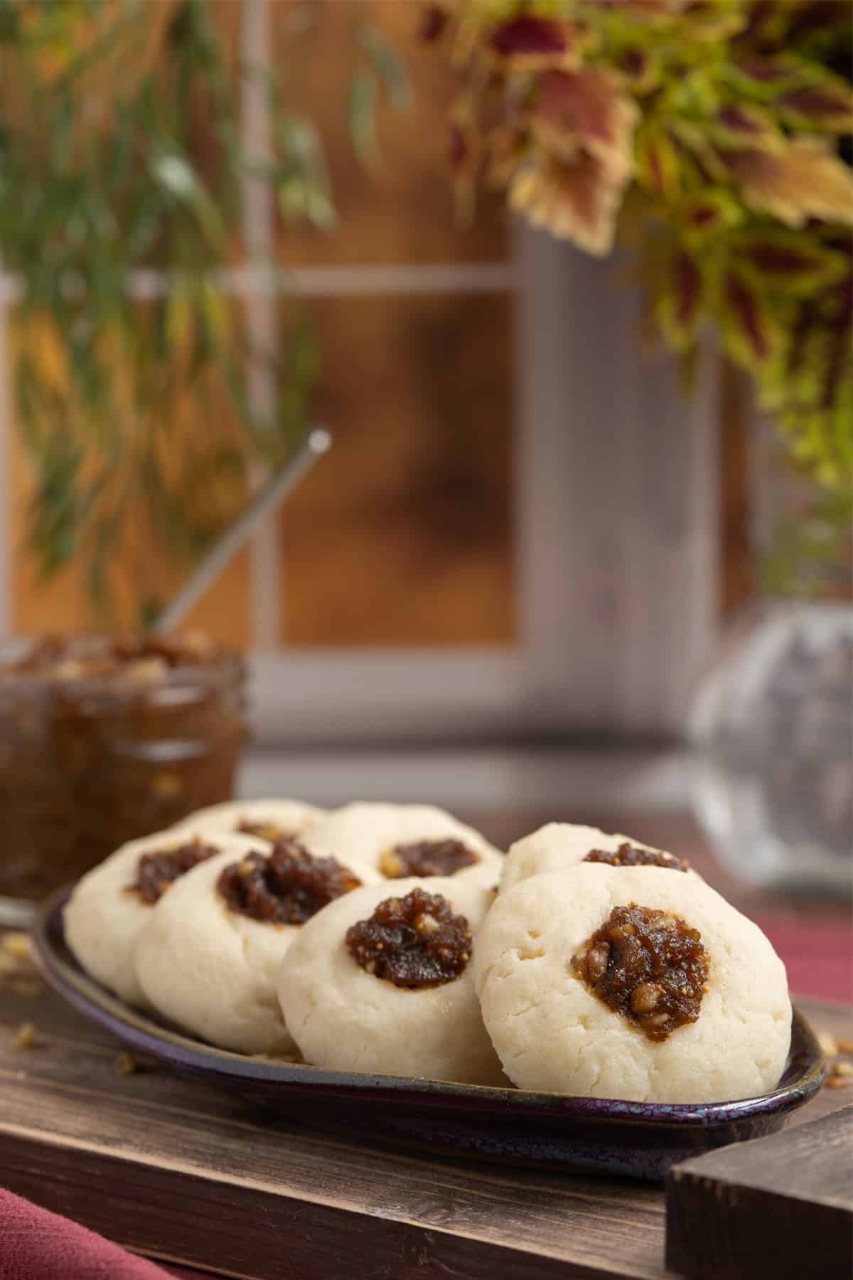 Orange fig thumbprint cookies on an oval dish sitting on a wooden plank in front of a window.
