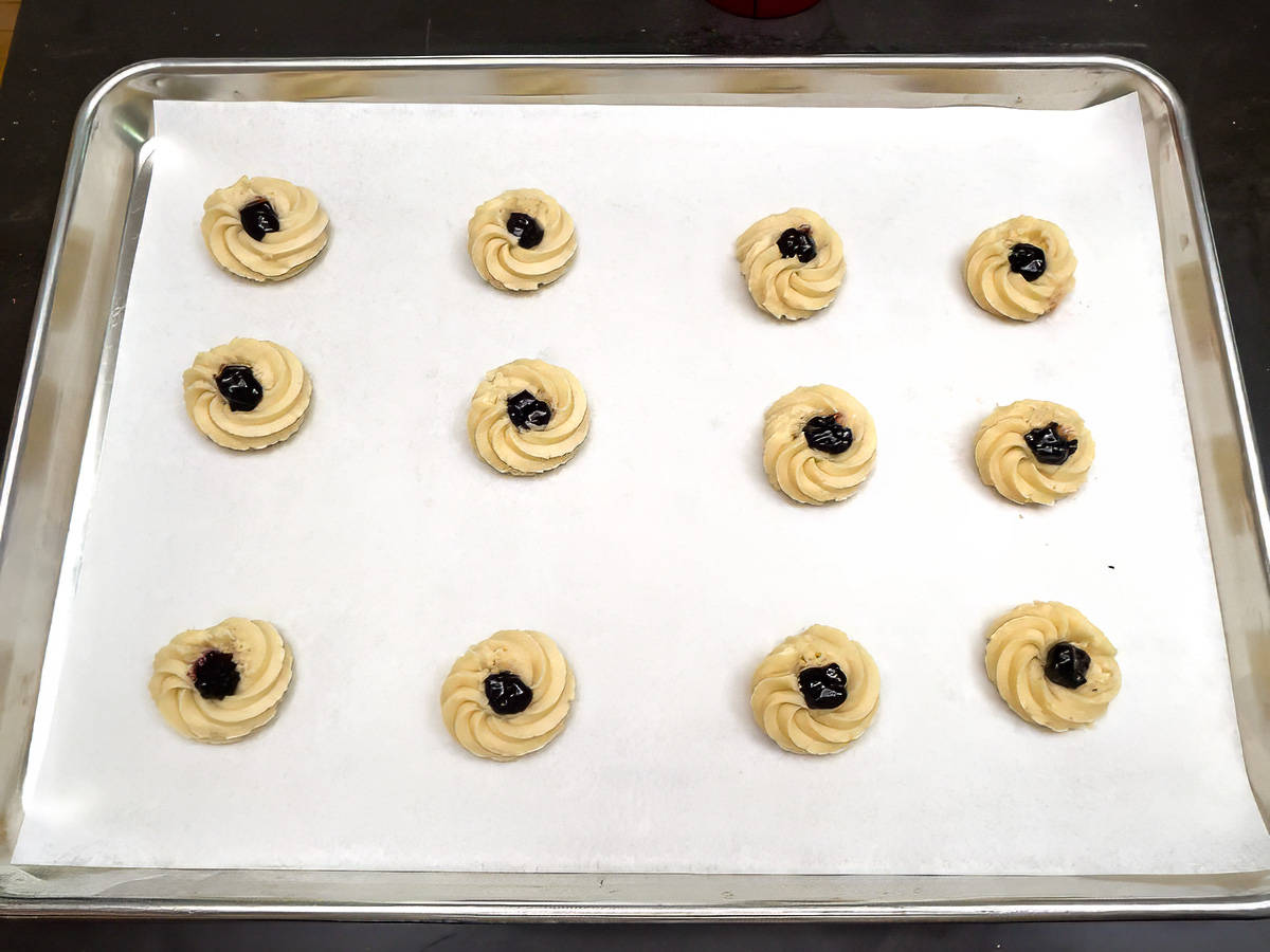 Twelve rosette cookies filled with jam in the middle, ready for the oven on a parchment-lined sheet pan.