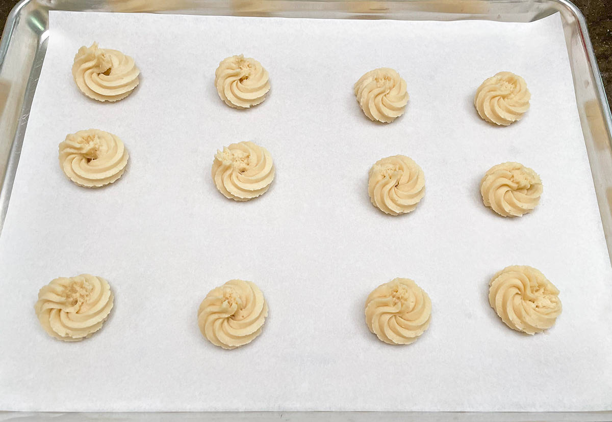 Twelve cookie dough rosettes after being piped onto a parchment-lined cookie sheet pan.