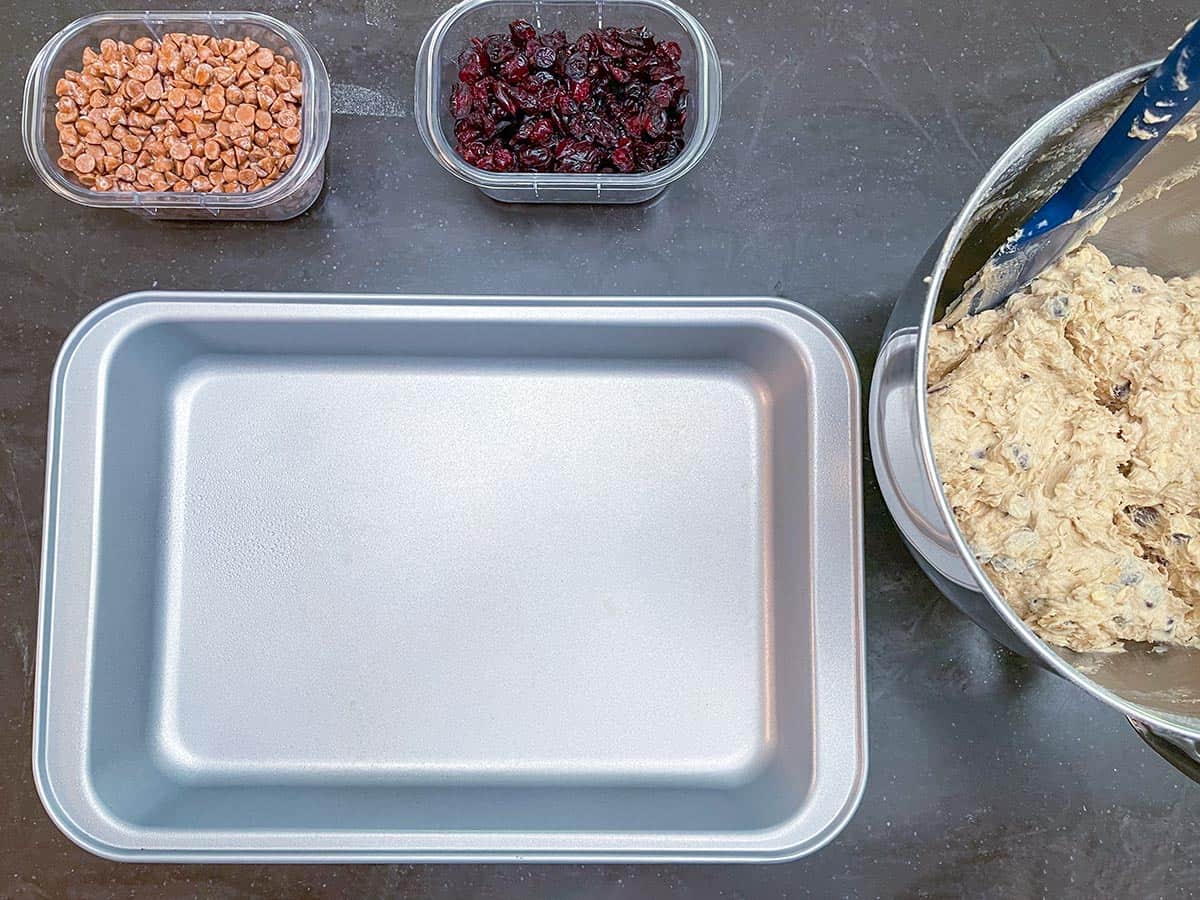 Set up for assembling, cinnamon chips, dried cranberries, mixer bowl with the cookie dough and the pan for baking.