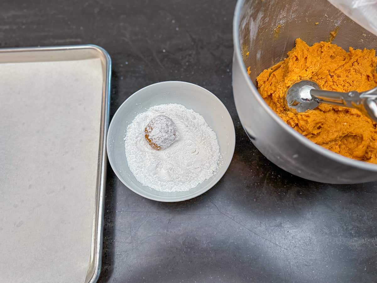 A bowl of the pumpkin cookie dough next to a bowl of powdered sugar and sitting next to a parchment-lined sheet pan.