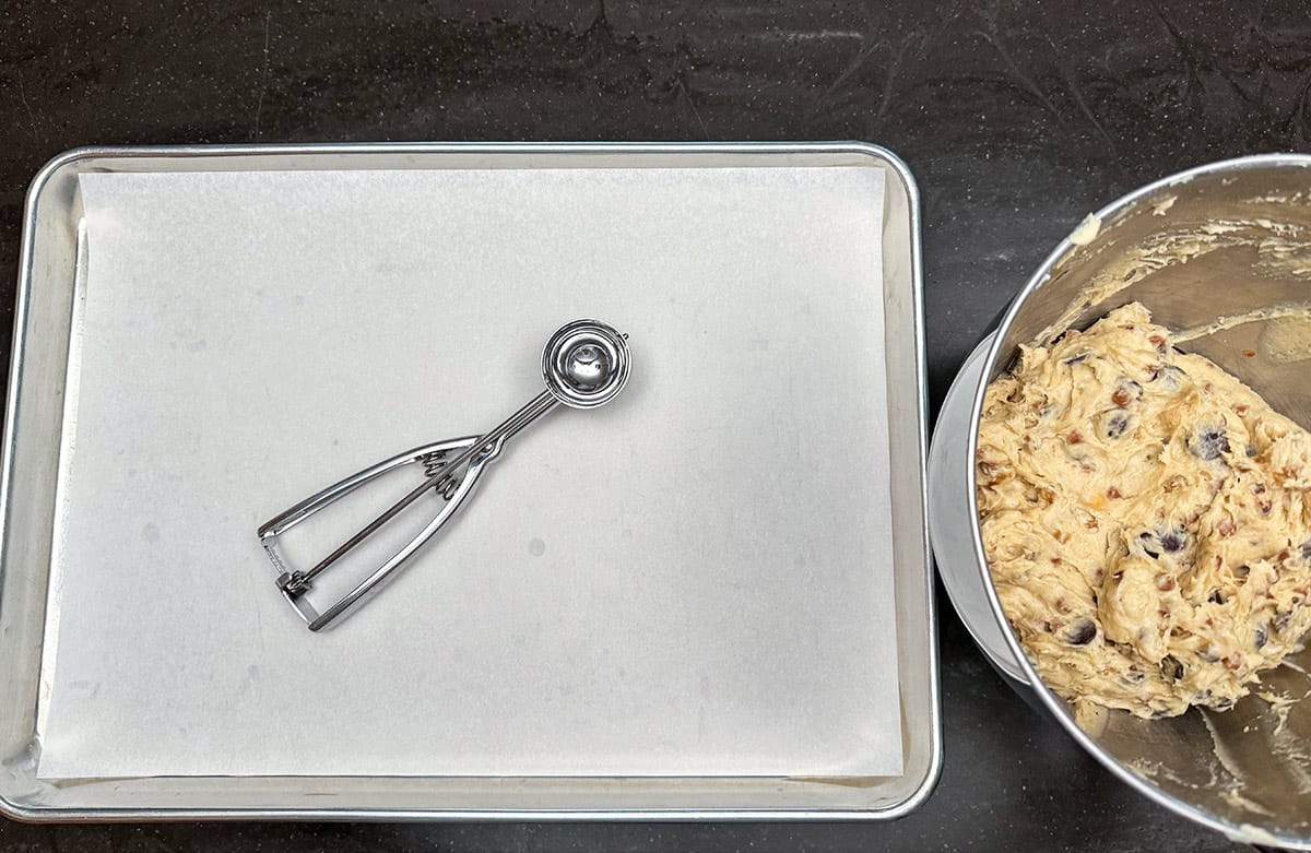 Medium cookie scooper on a parchment-lined sheet pan. The bowl of cookie dough sitting beside the pan.