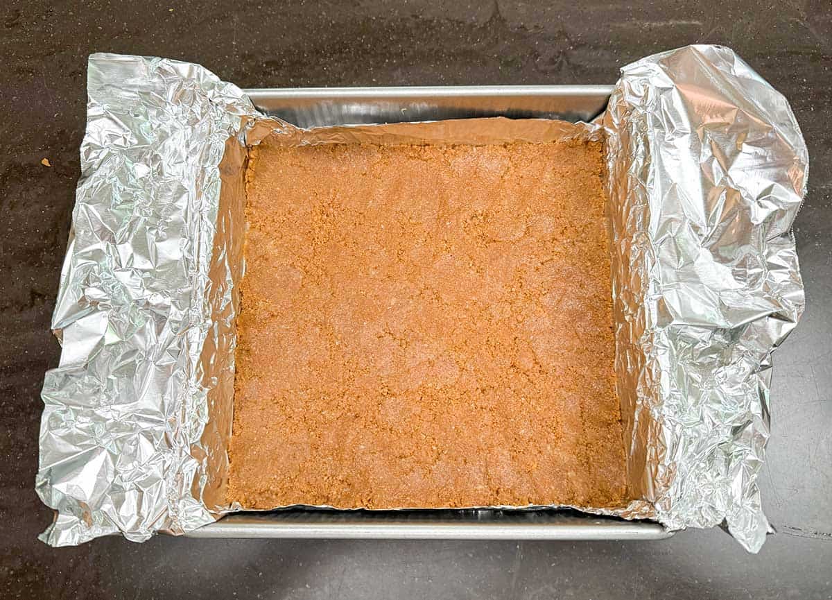 The crust has been pressed along the bottom of the tin foil-lined baking pan.
