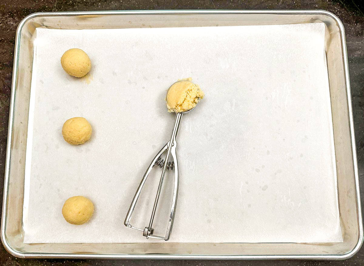 Sheet pan with three cookie dough balls along with a scooper.