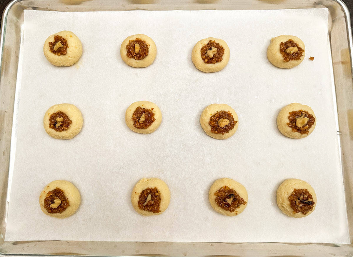 Cookies filled with the fig filling and a walnut on top ready for the oven on a cookie sheet pan.