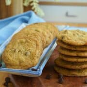 A stack and a tray of Thin and crispy chocolate chip cookies on a wooden plank.