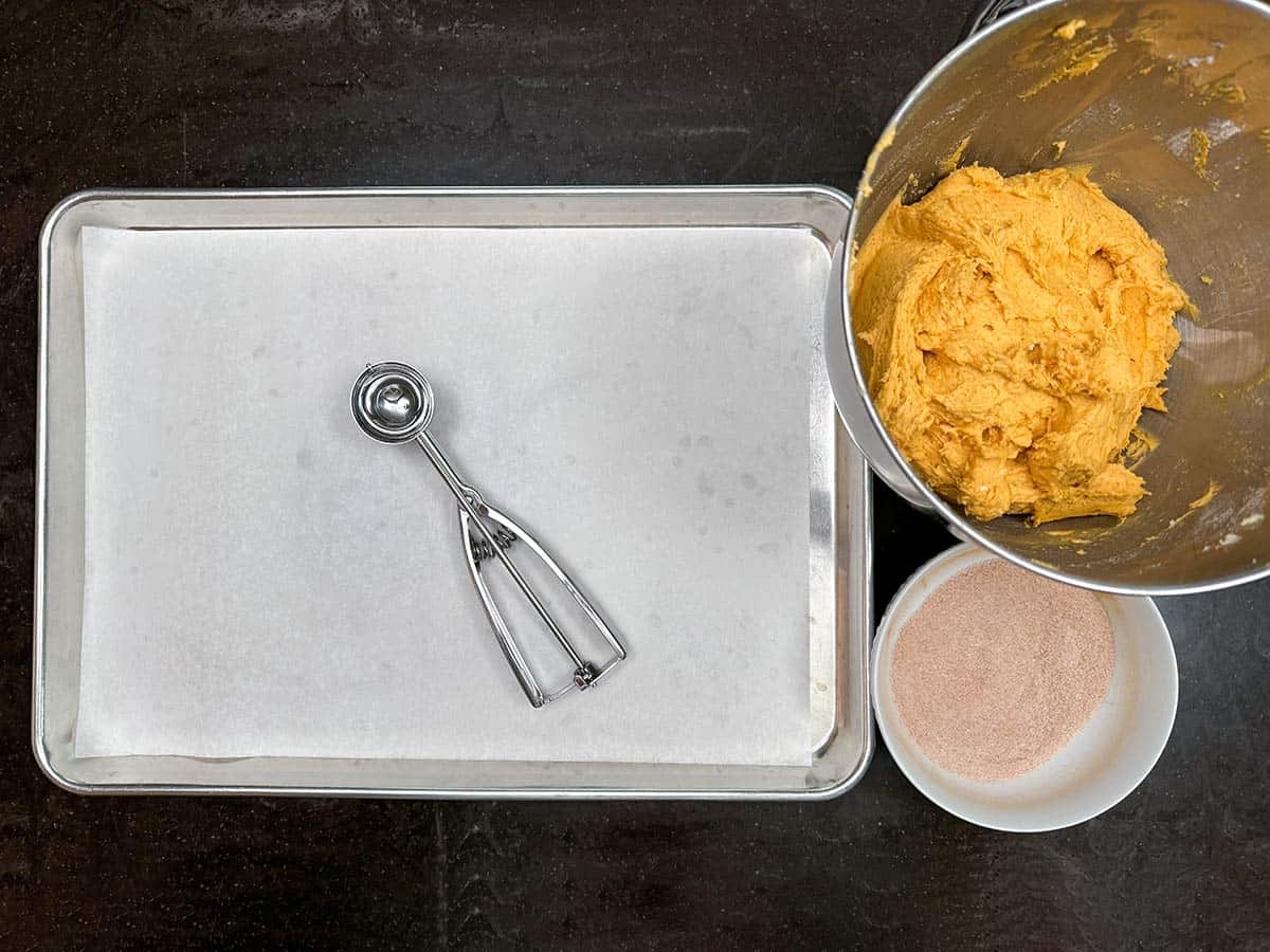 A bowl with the cookie dough, a cookie scooper on a parchment lined sheet pan, along with a bowl of the sugar-cinnamon mix ready for assembling.