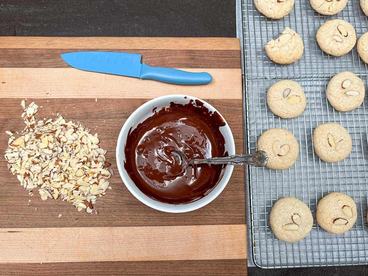 On a cutting board chopped almonds and the bowl of melted chocolate next to the cooled cookies to finish the topping.