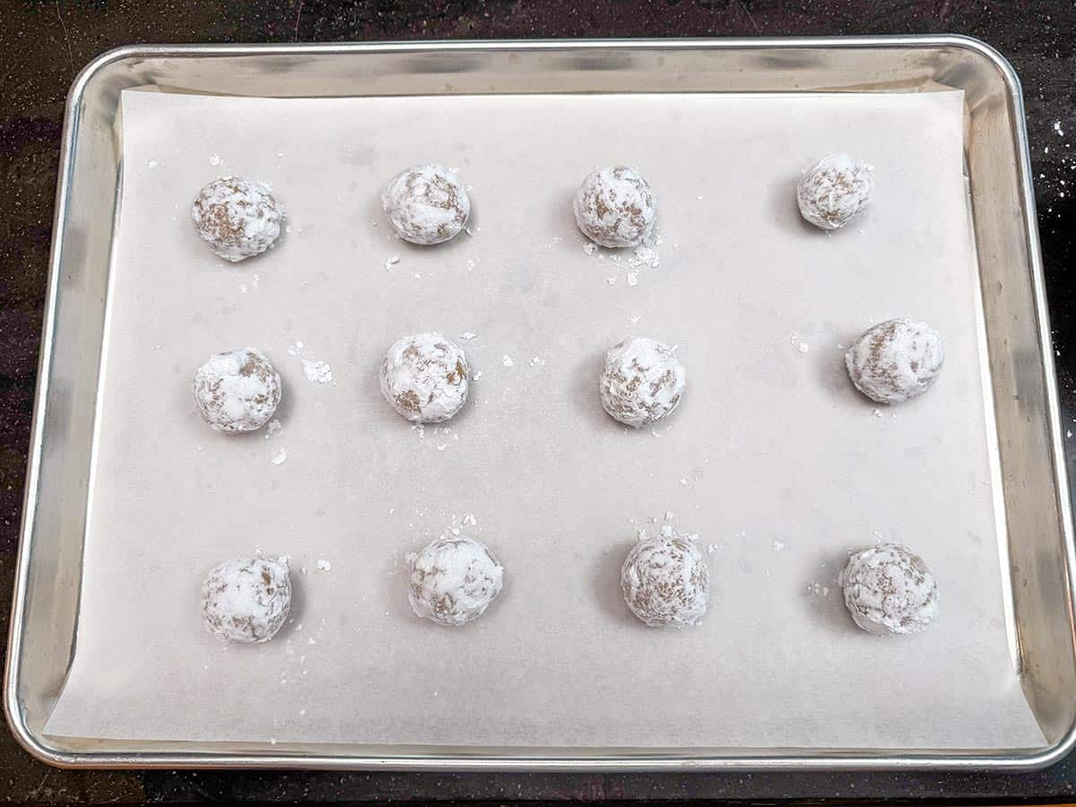 Cookie balls that are coating with powdered sugar sitting on a parchment lined cookie sheet pan.