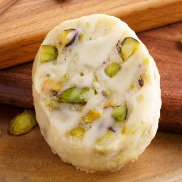 A single front view of a lemon pistachio cookie leaning against a wooden board.
