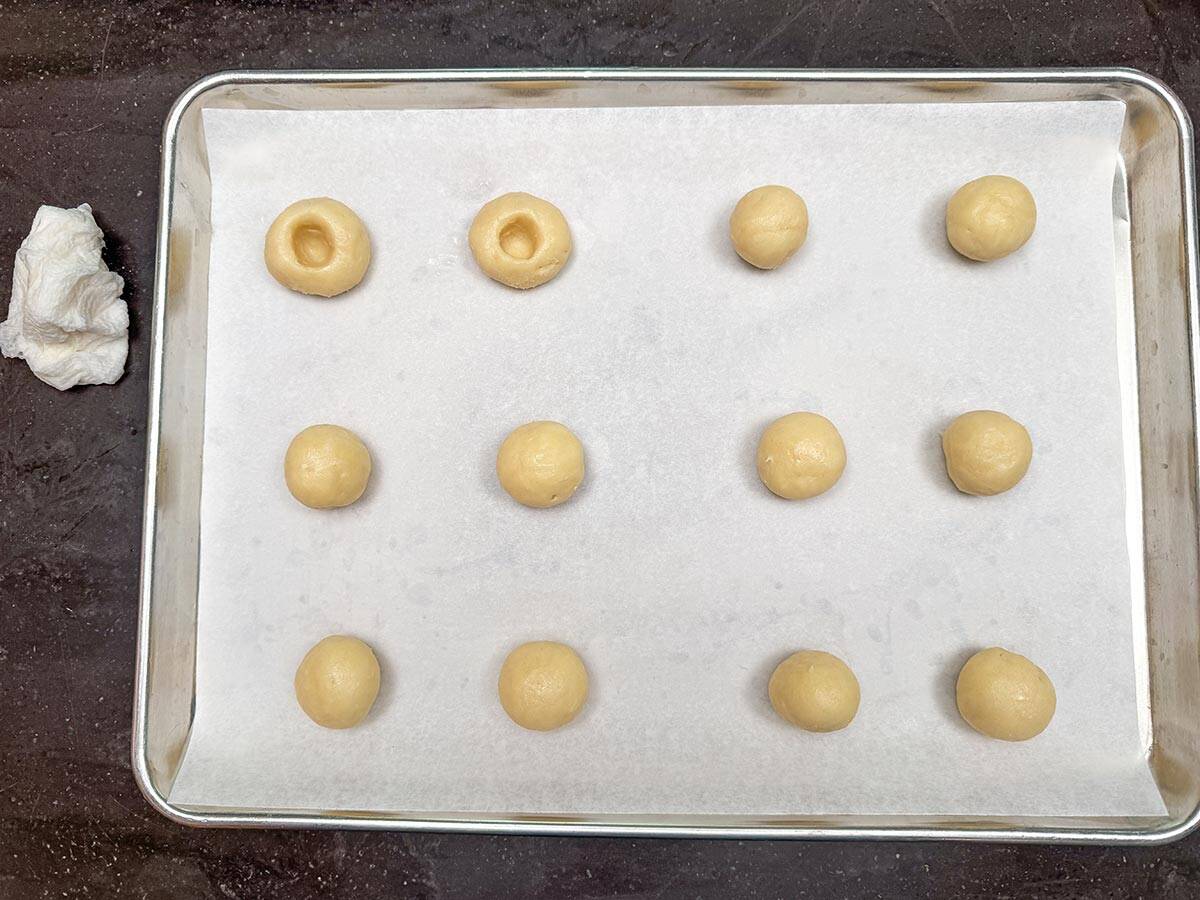 Twelve cookie balls on a sheet pan with two of the balls with thumb imprint.