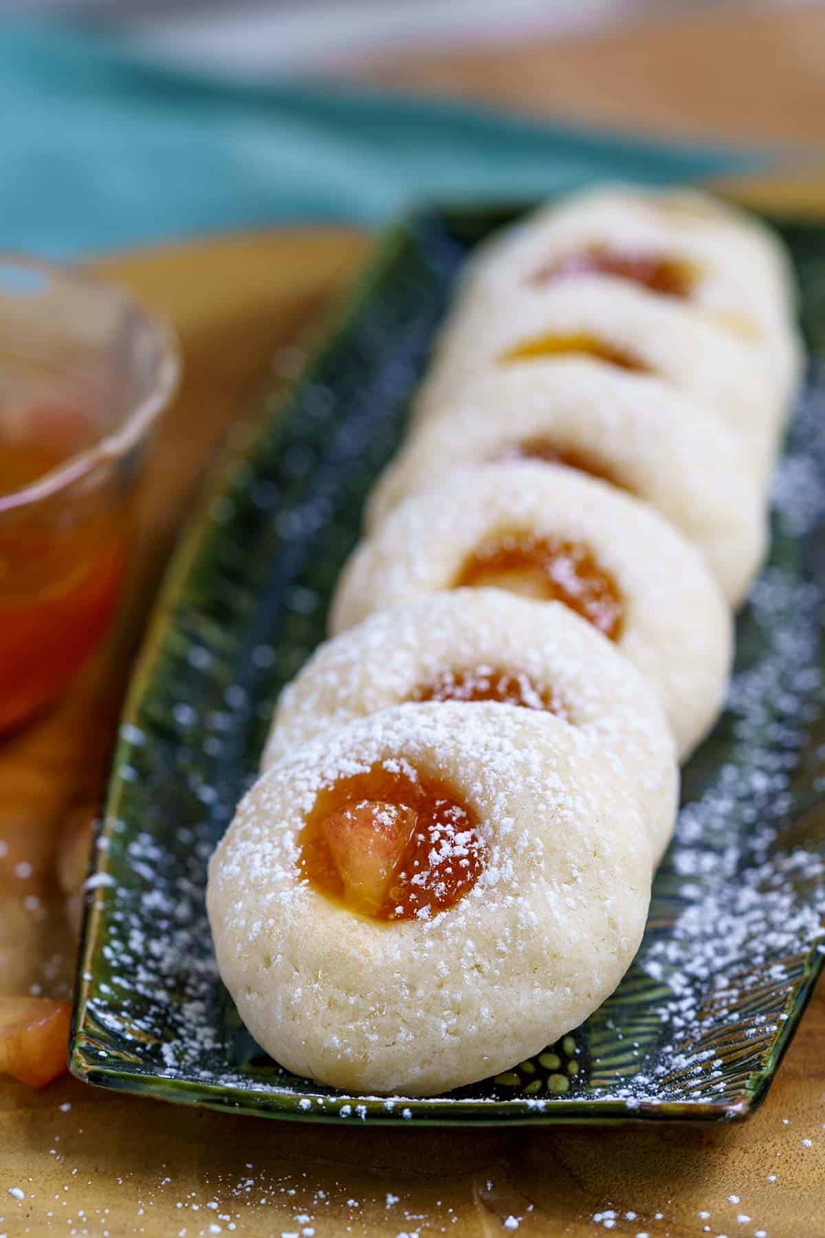 Fresh peach and preserve thumbprint cookies lined up on a narrow plate with powdered sugar sprinkled on top.