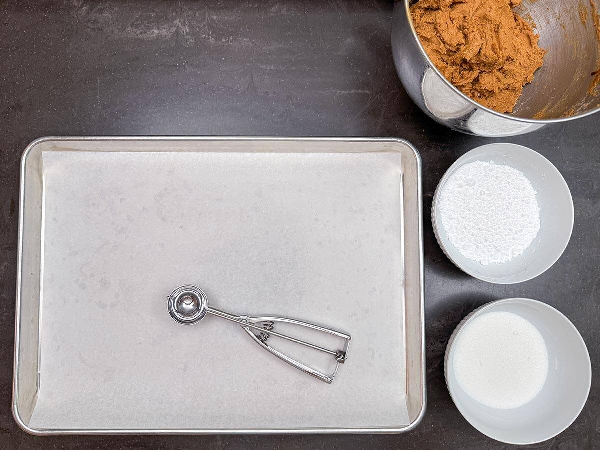 Cookie sheet pan along with a cookie scooper, also two bowls of sugar for coating cookie balls.