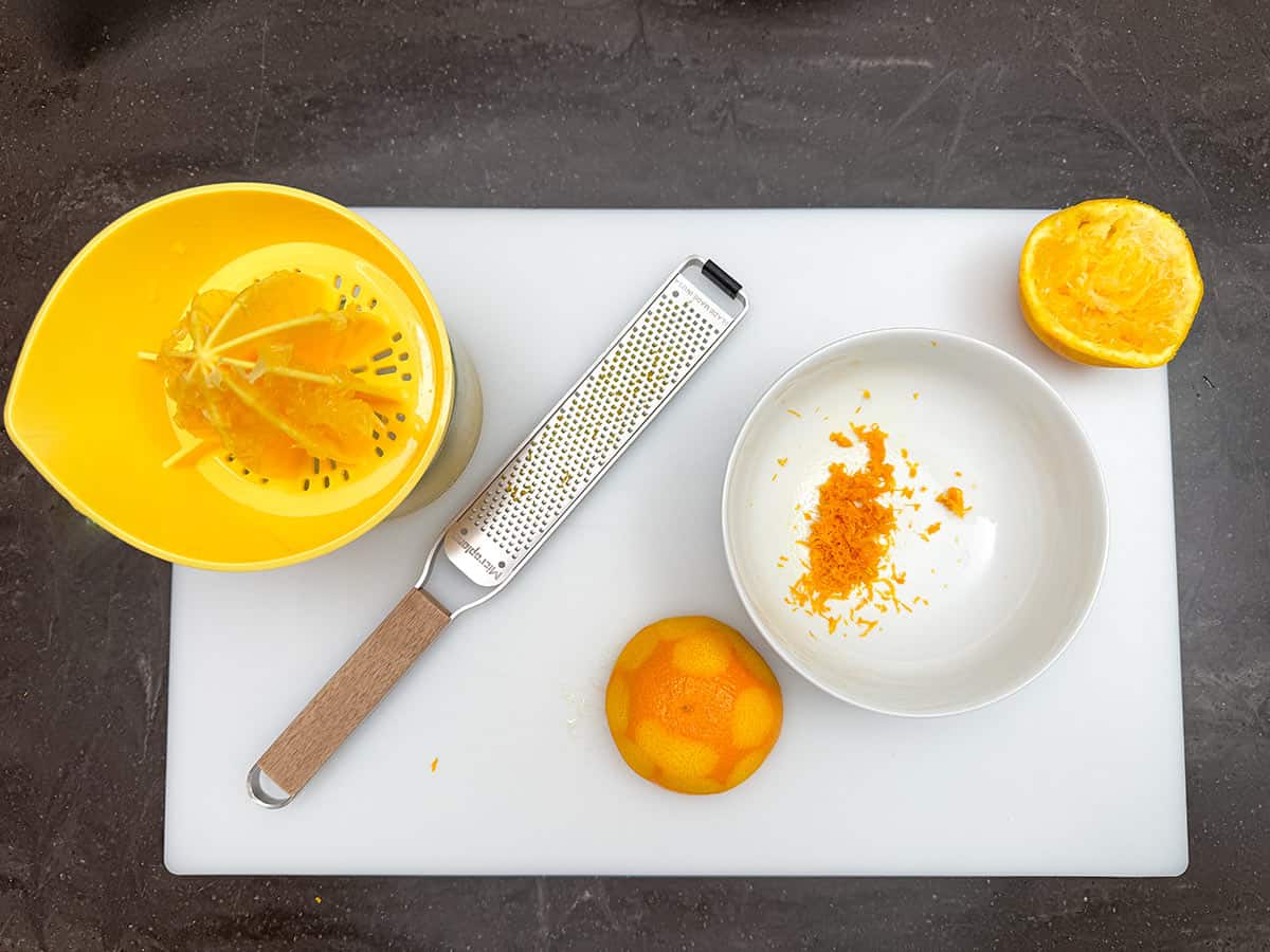 A bowl, juicer and a zester on a cutting board with an orange.