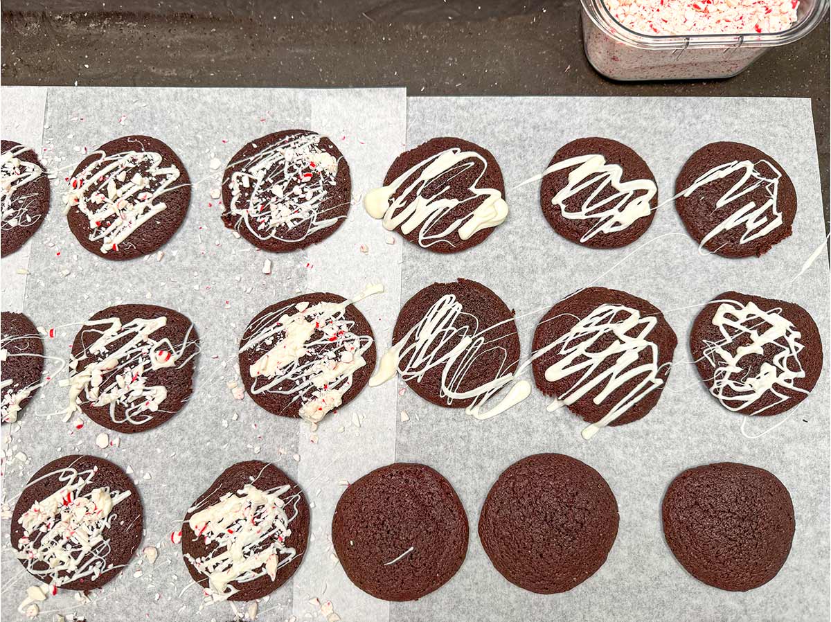 Baked cookies on parchment paper getting the white chocolate drizzle and peppermint pieces on top.