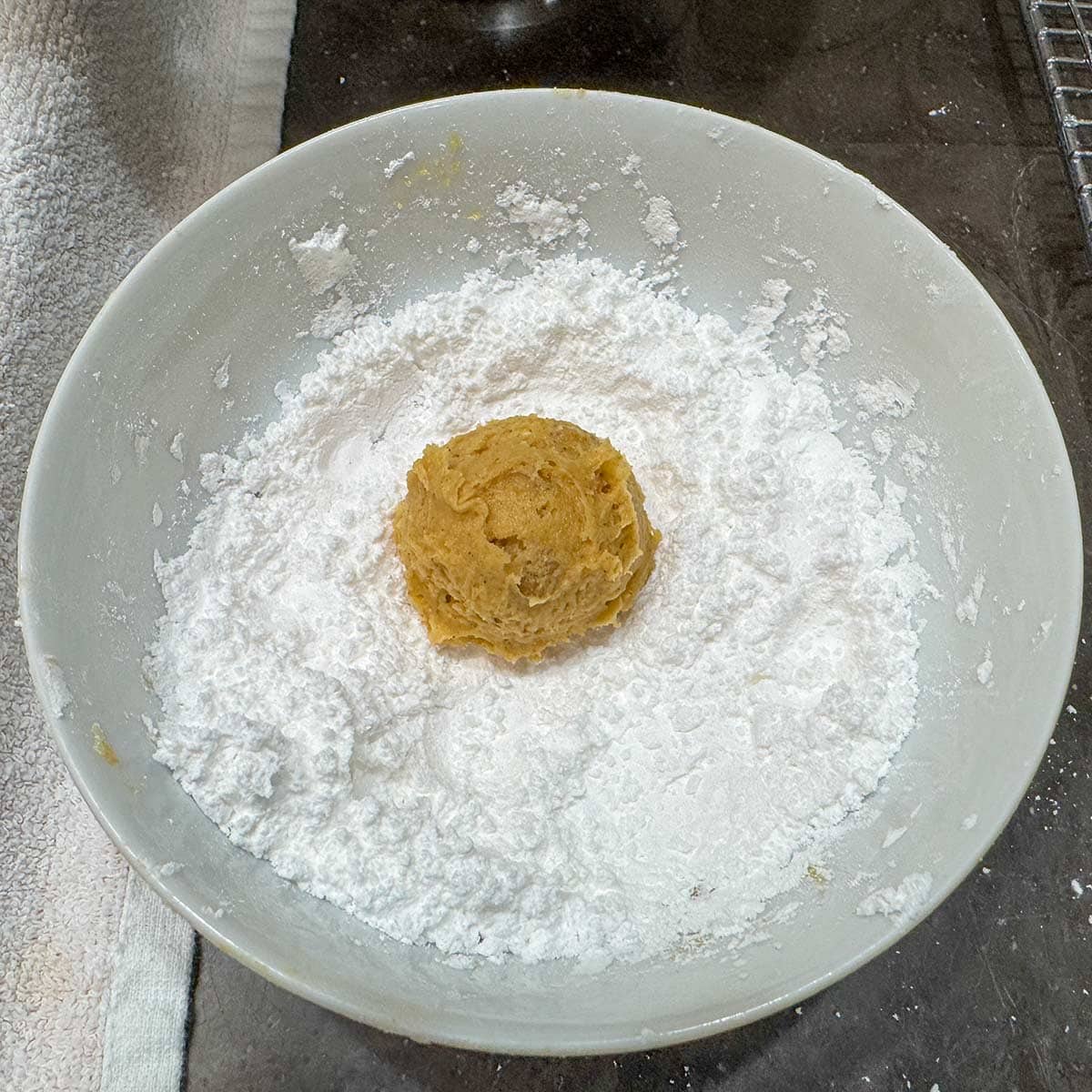 A scooped ball of cookie dough added to a bowl of powdered sugar ready to be coated.