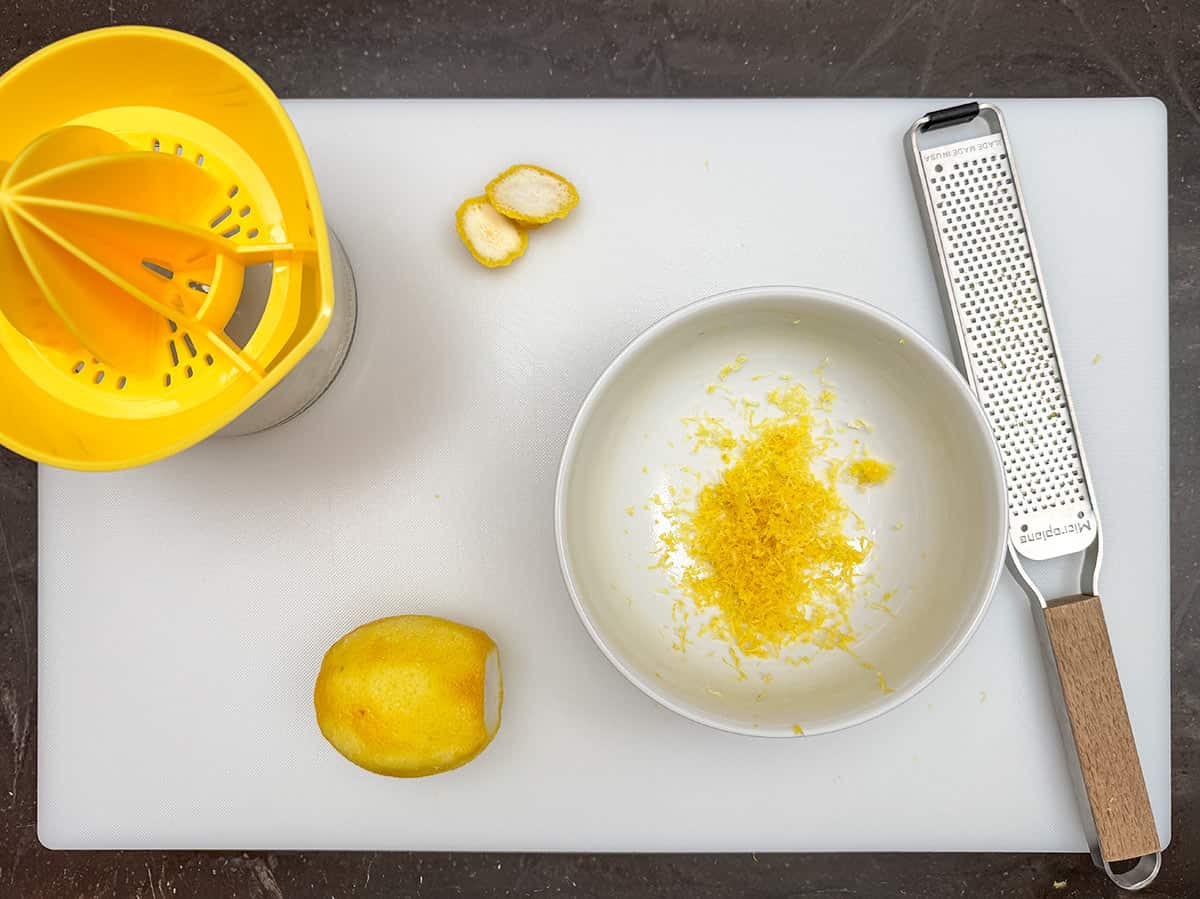 Lemon zest in a bowl and the lemon will be cut in half to be juiced.