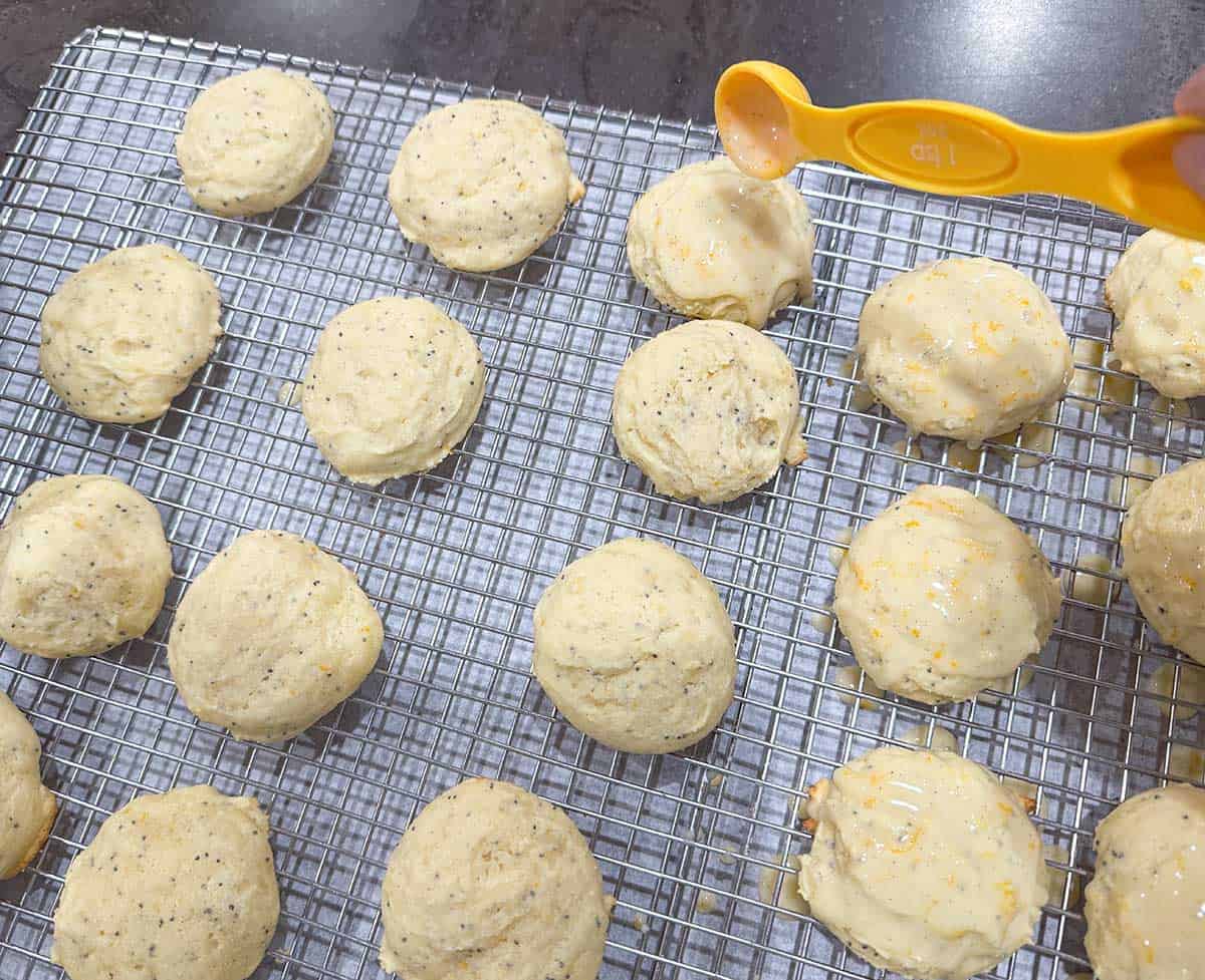 Using a teaspoon to add the glaze onto the tops of the cooled cookies that are on a wire rack.
