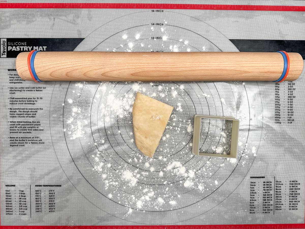Pastry mat with a quarter of the dough, square cookie cutter, and a wooden dowel with bands at the ends for even rolling.
