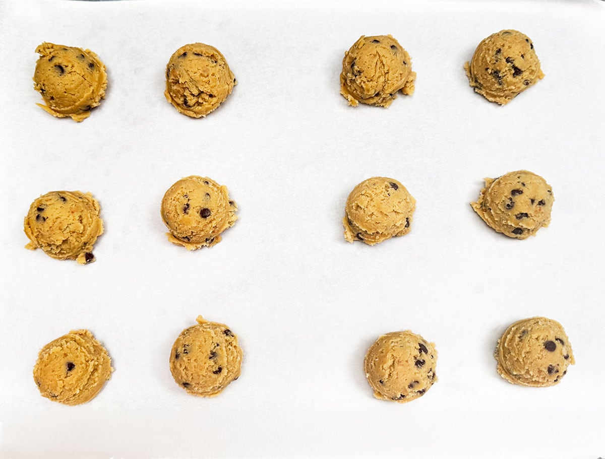 Twelve scoops of cookie dough on a parchment-lined cookie sheet pan.