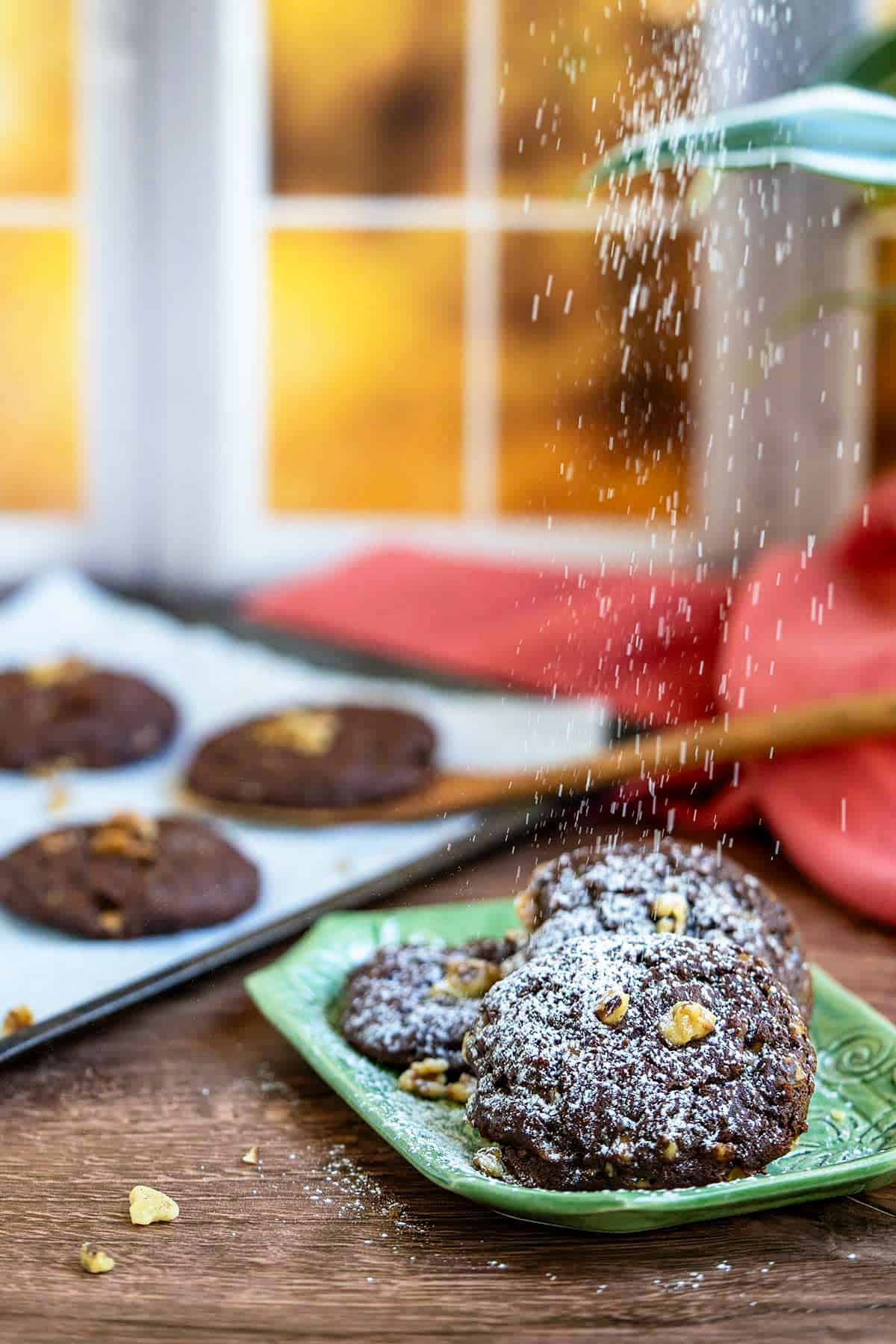 Finished chocolate orange toffee cookies on a square plate with powdered sugar falling on top of the cookies.