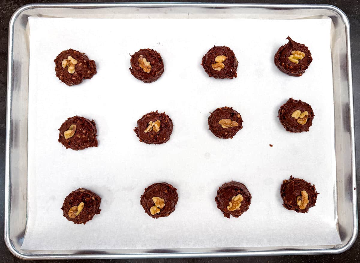 Scooped cookies with a couple of walnuts on top sitting on a parchment-lined sheet pan.