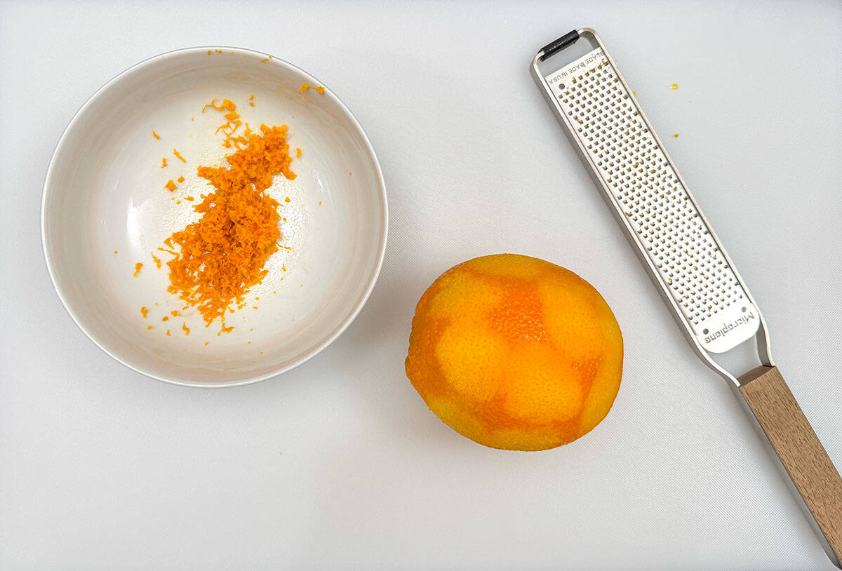 A bowl with orange zest added along with the orange and the zester.