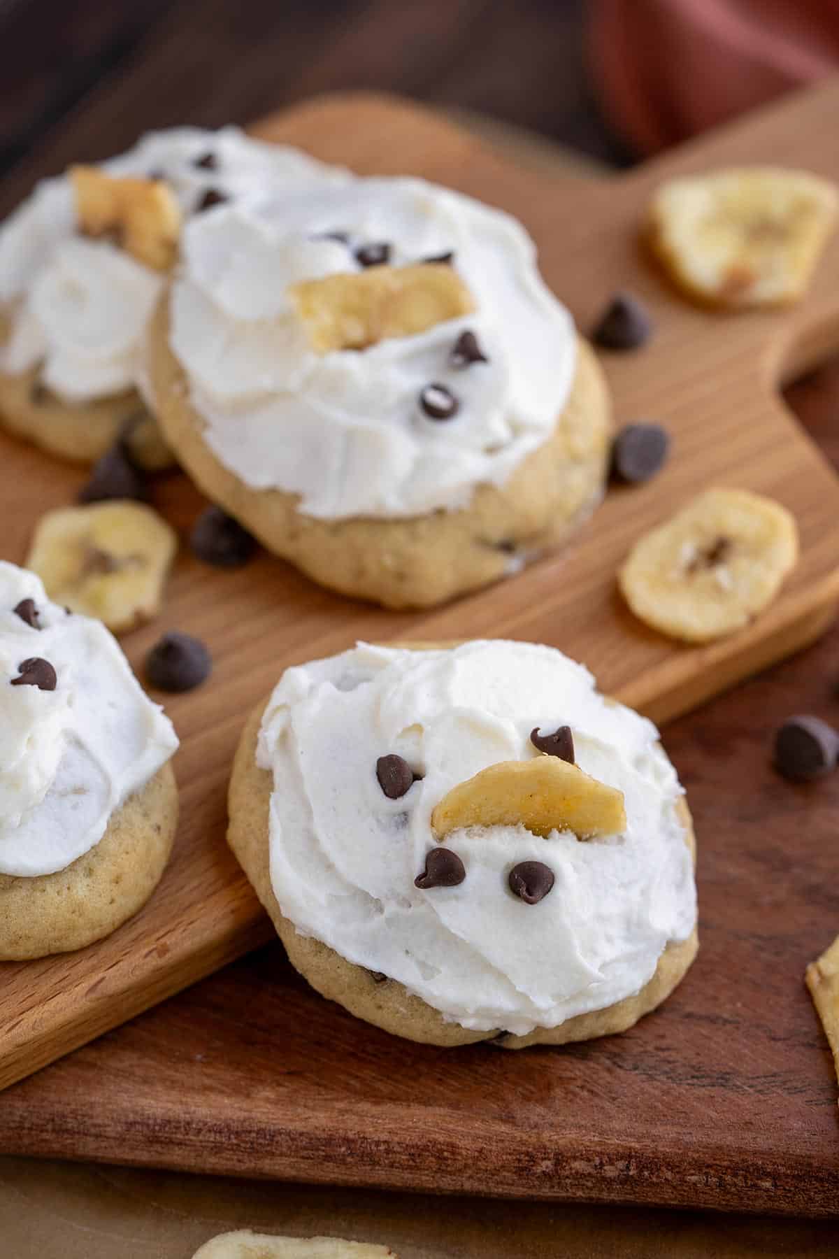 Banana Bread cookies with mini chocolate chips and a banana chip on top of the icing. sitting on a wooden board.