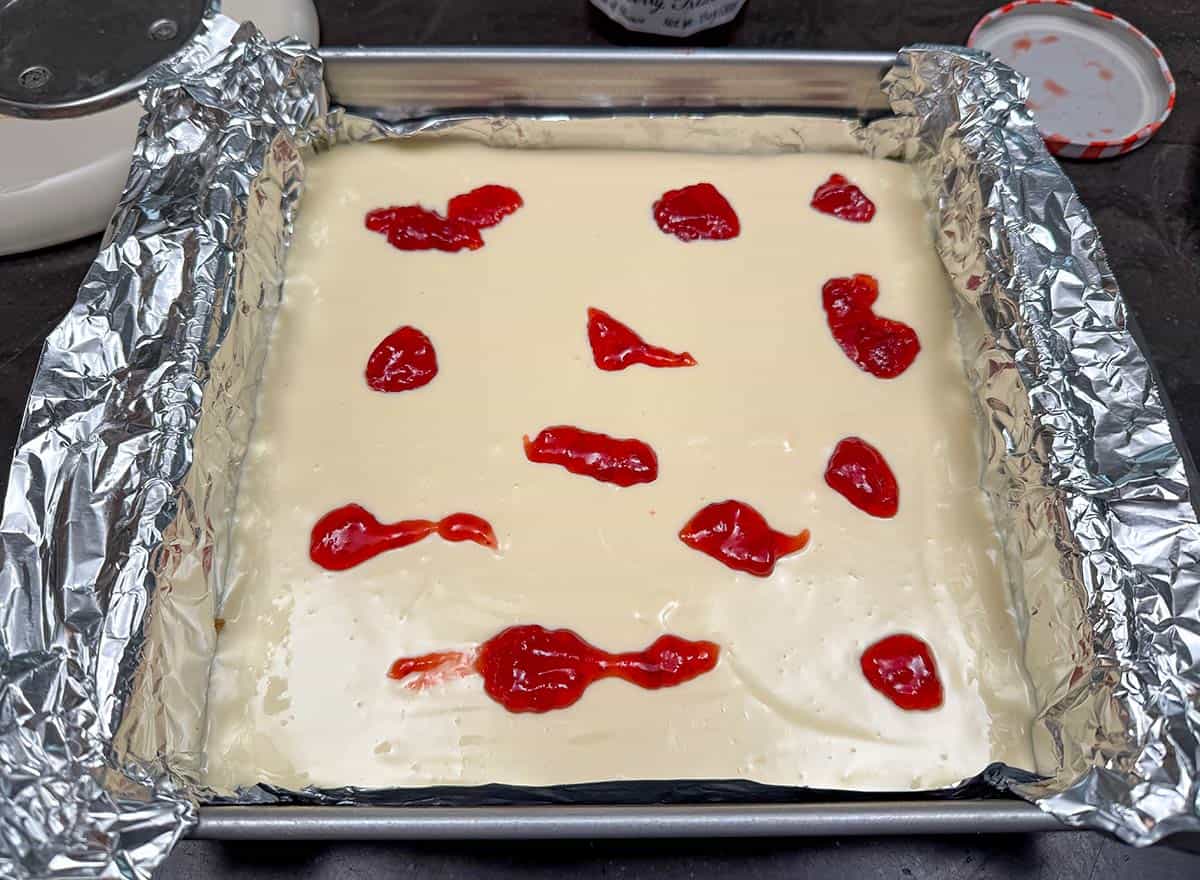 Unbaked cheesecake getting dollops of strawberry preserves around the top.