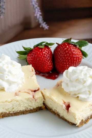 Two strawberry key lime cheesecake squares with whip cream and two strawberries behind the cheesecake pieces.