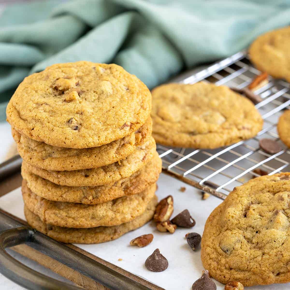 Chocolate chip cookies with glazed pecans stacked on a tray with nuts and chocolate chips laying around.