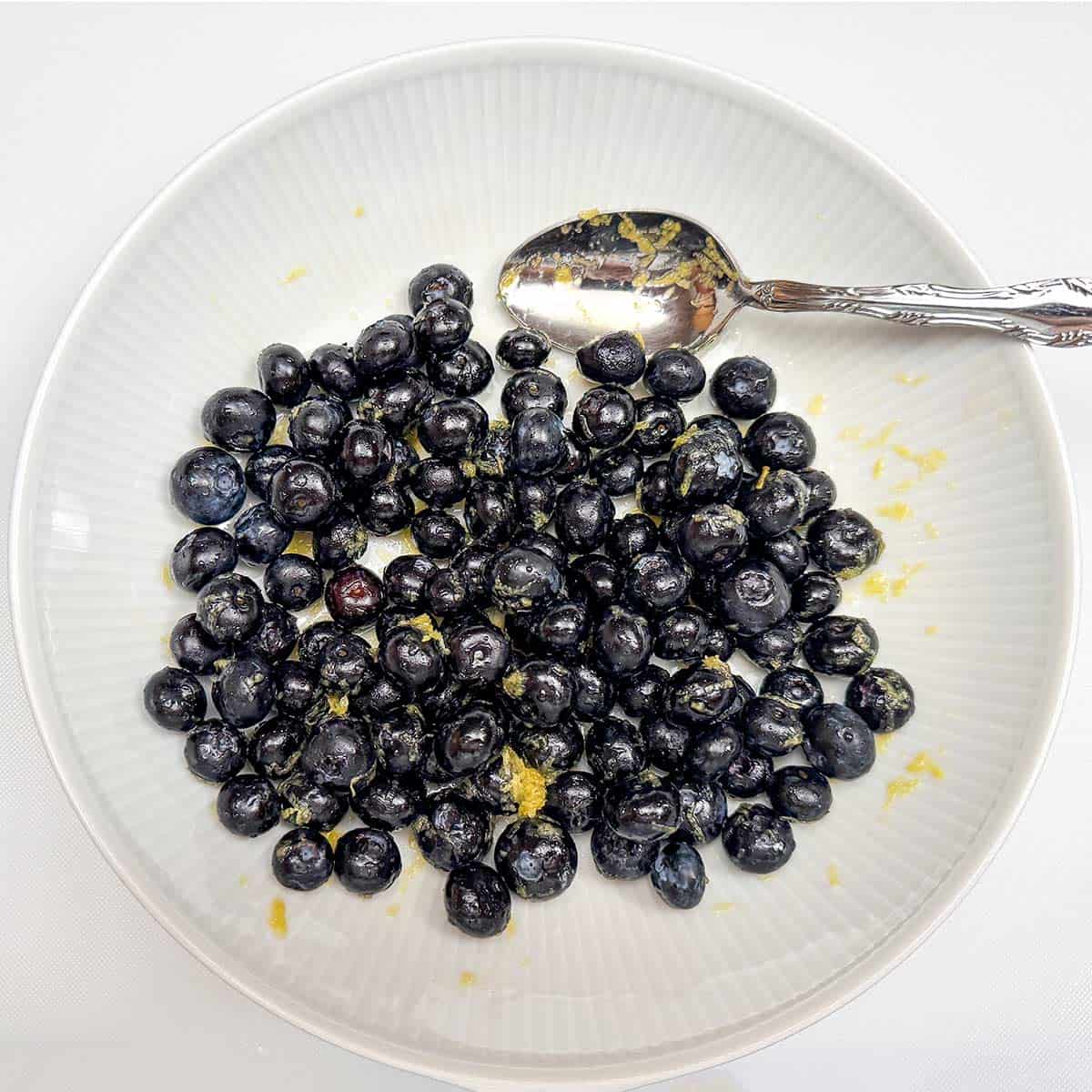 Infusing whole fresh blueberries in a bowl with lemon zest and maple syrup.