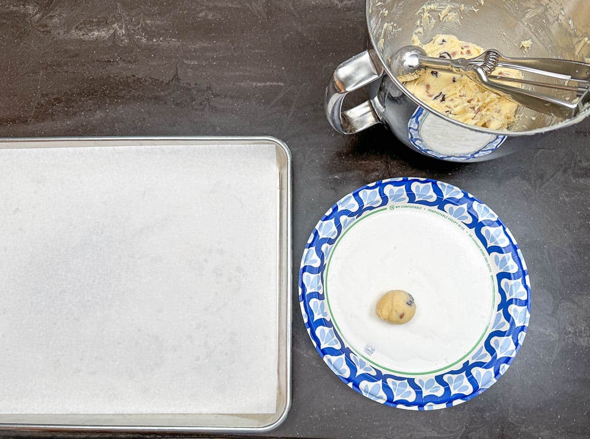 Rolling and coating a ball of cookie dough into fine sugar to be placed on a parchment-lined cookie sheet pan.
