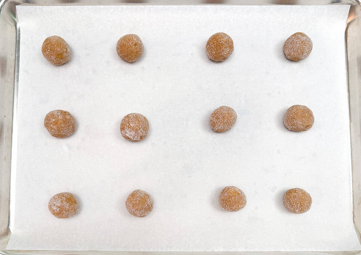 Twelve cookie balls that have been rolled in sugar on a parchment paper lined sheet pan.