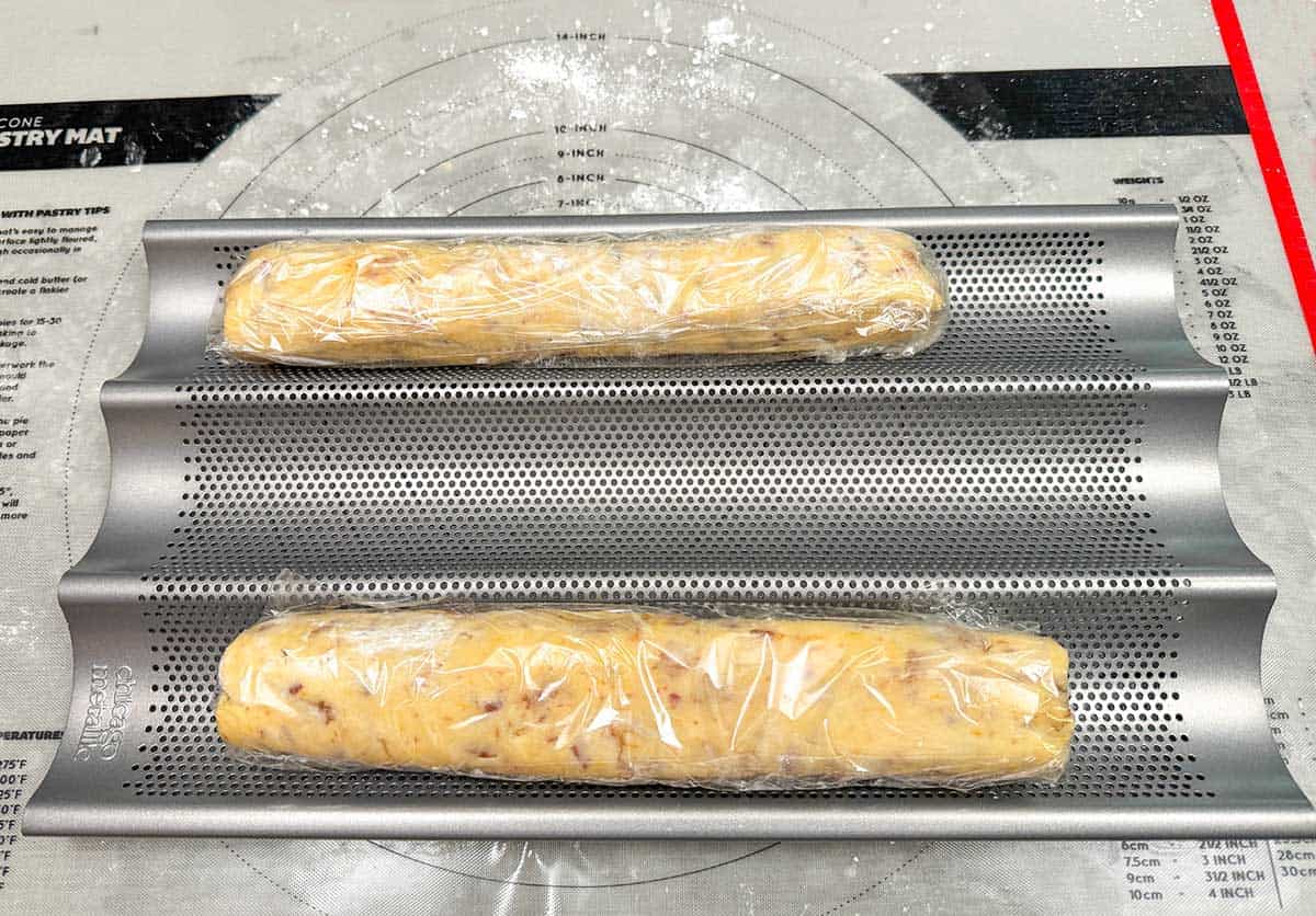 Two cookie logs that have been wrapped in plastic wrap and sitting in a baguette pan.