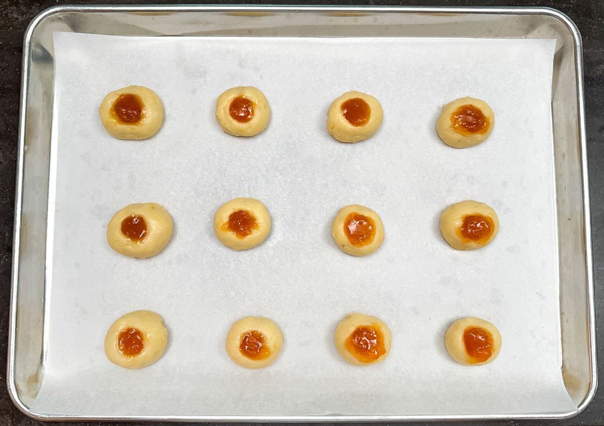 Cookie balls that have the holes added and filled with apricot preserves on a parchment-lined sheet pan.