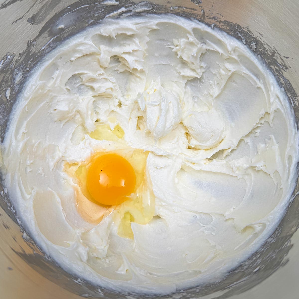 An egg added to a bowl of cream cheese, butter and sugar mixture.
