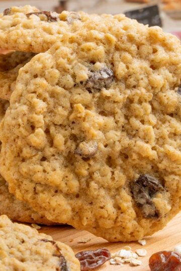 A stack and front view of Spiced Oatmeal Raisin Cookies on a board with oats and raisins around the cookies.