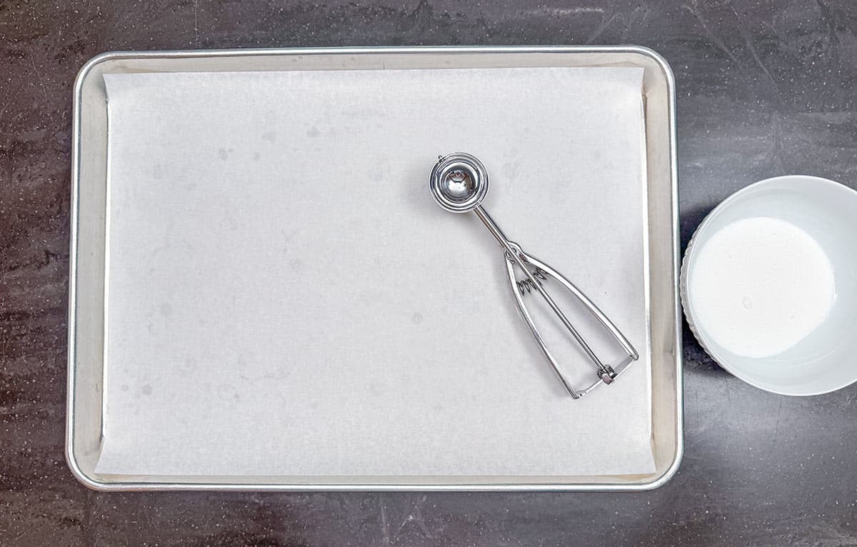 A bowl with superfine sugar beside a parchment paper lined sheet pan with a medium cookie scoop laying on top.