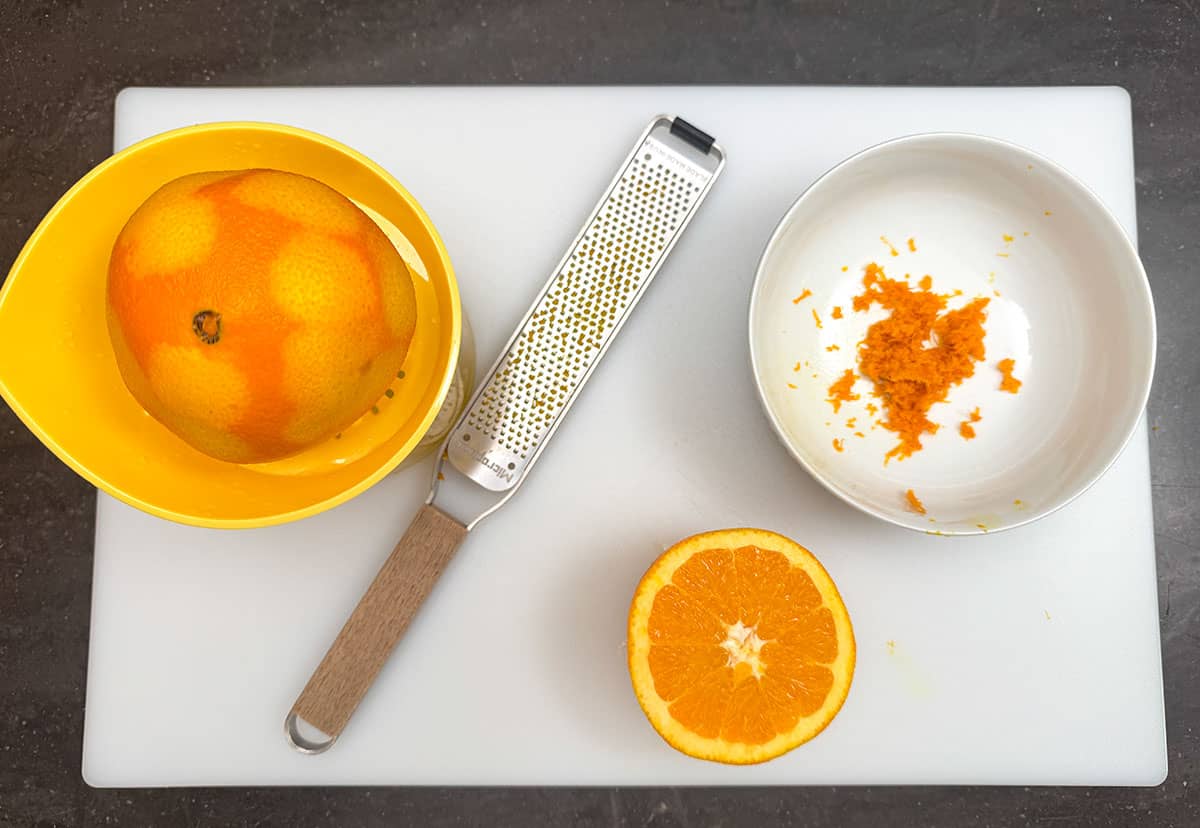 Zesting the whole orange into a bowl and juicing half oranges with a manual juicer on a cutting board.