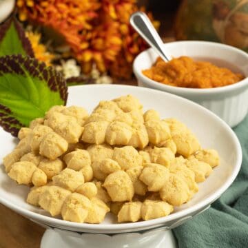 A plate full of finished Pumpkin Pie Spritz Cookies with a small bowl of pumpkin puree and spoon.