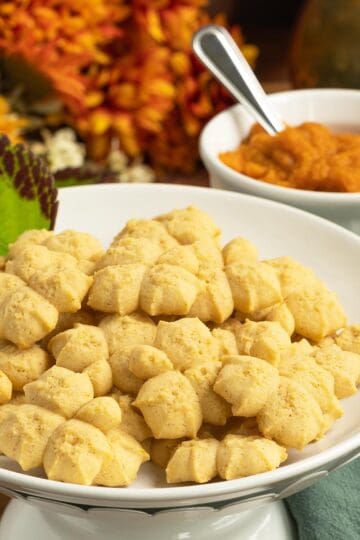 A plate full of finished Pumpkin Pie Spritz Cookies with a small bowl of pumpkin puree and spoon.